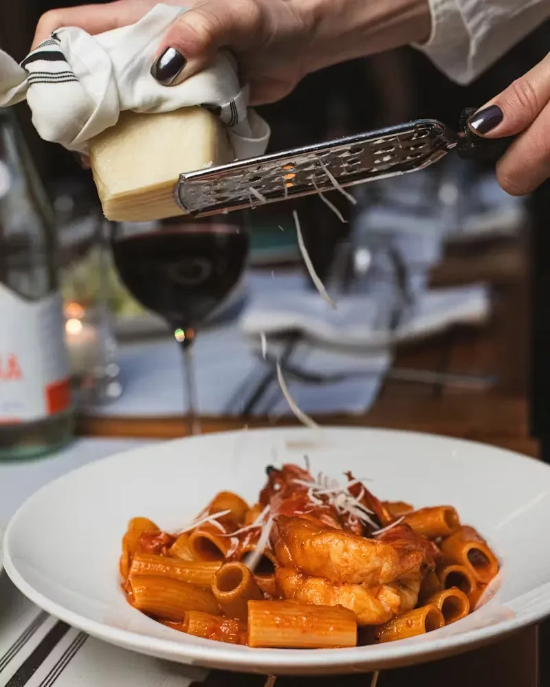 Hands grate cheese onto a plate of rigatoni pasta with shrimp.
