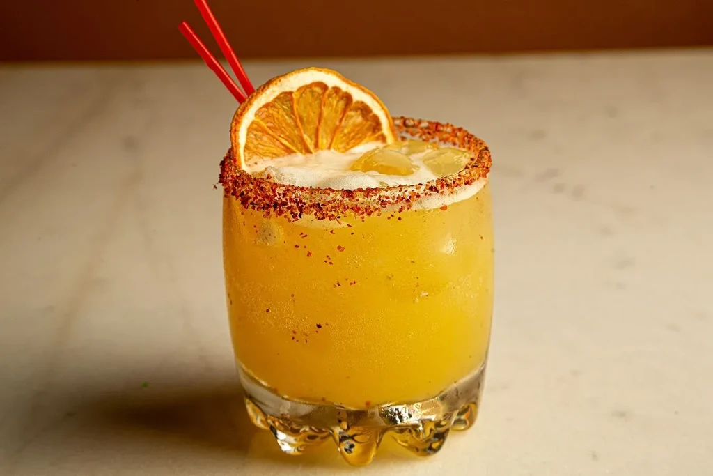 A glass of yellow cocktail with ice and a dried orange slice.