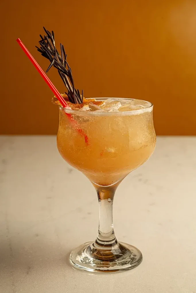 A chilled orange cocktail with rosemary and a red straw against an orange wall.