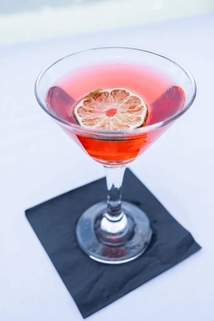 A martini glass with red liquid and a dried lime slice on a black napkin.