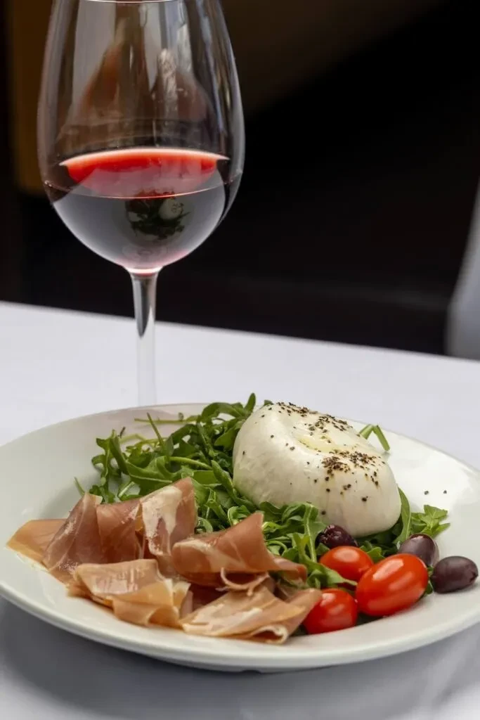 Red wine in a glass next to burrata, prosciutto, arugula salad.
