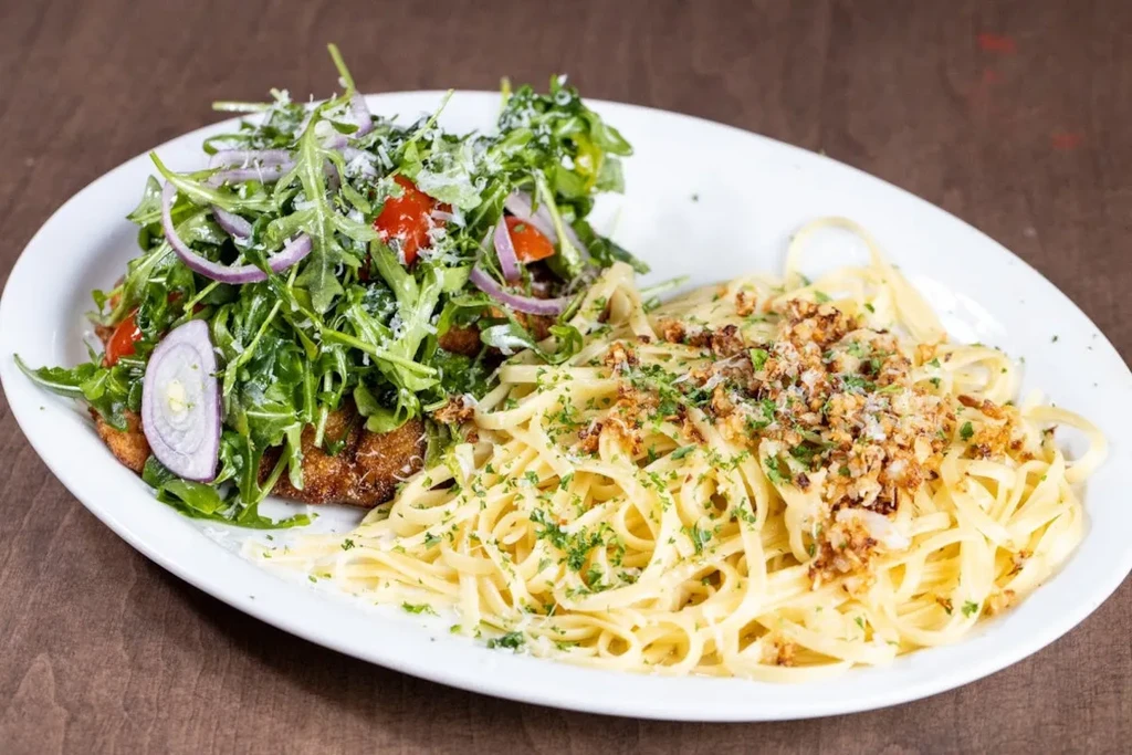 A white plate of pasta, chicken cutlet, and arugula salad on a wooden table.