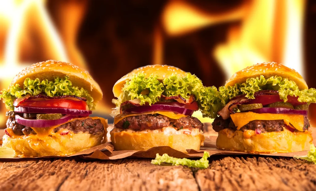 Three hamburgers on a wooden table, with flames in the background.