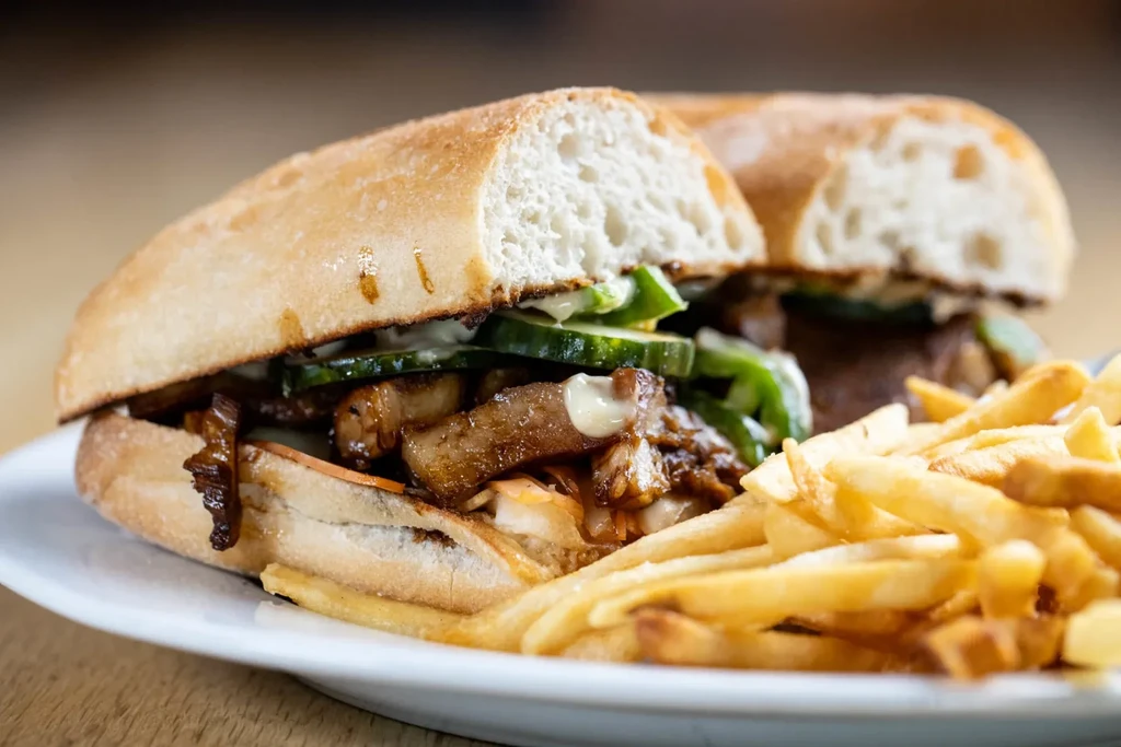 A pork sandwich with sliced cucumbers, sauce, and a side of fries.