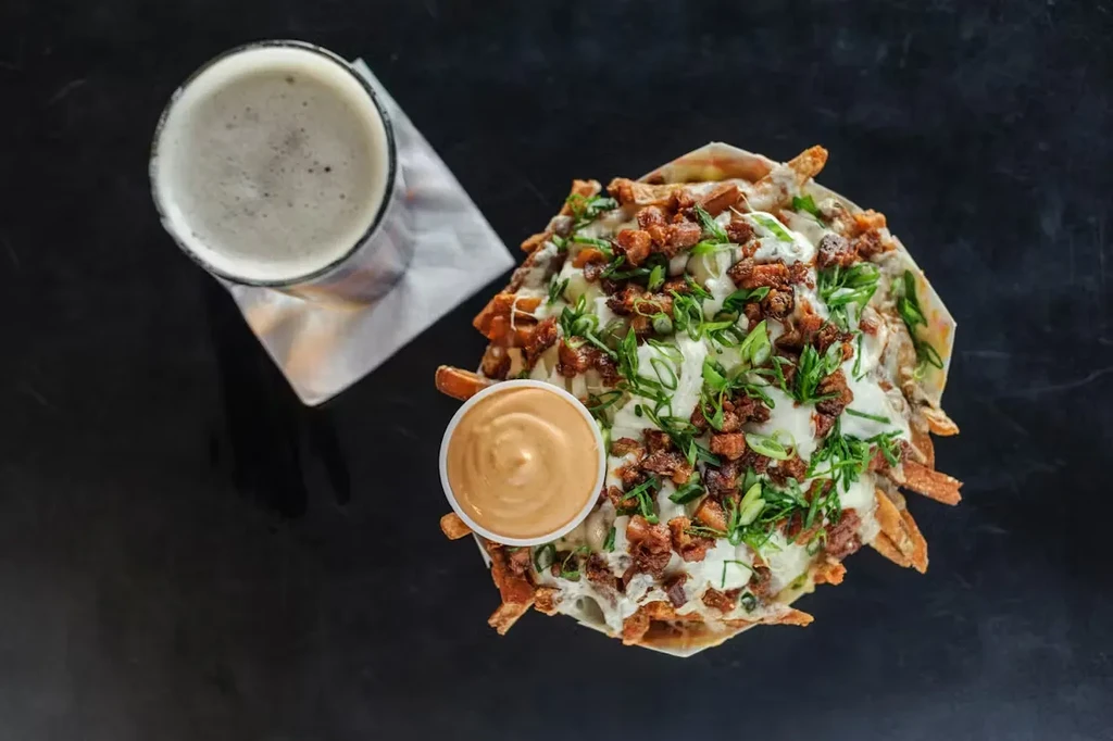 Overhead view of beer and loaded fries with sauce on a dark surface.
