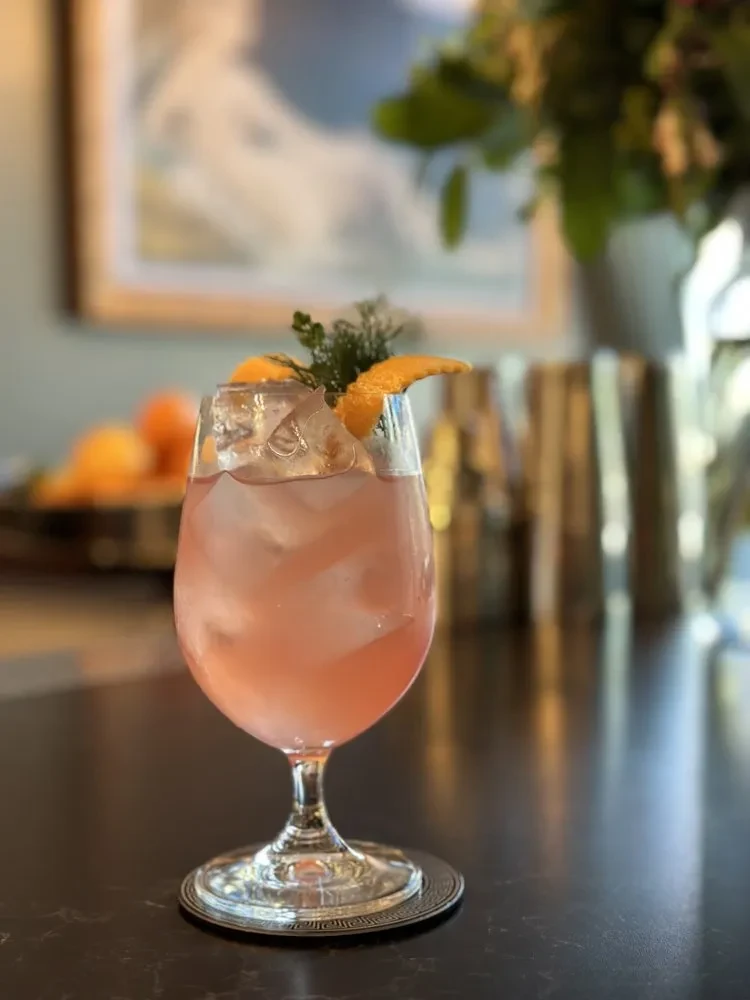 A pink cocktail with ice, orange peel, and herbs on a dark surface.