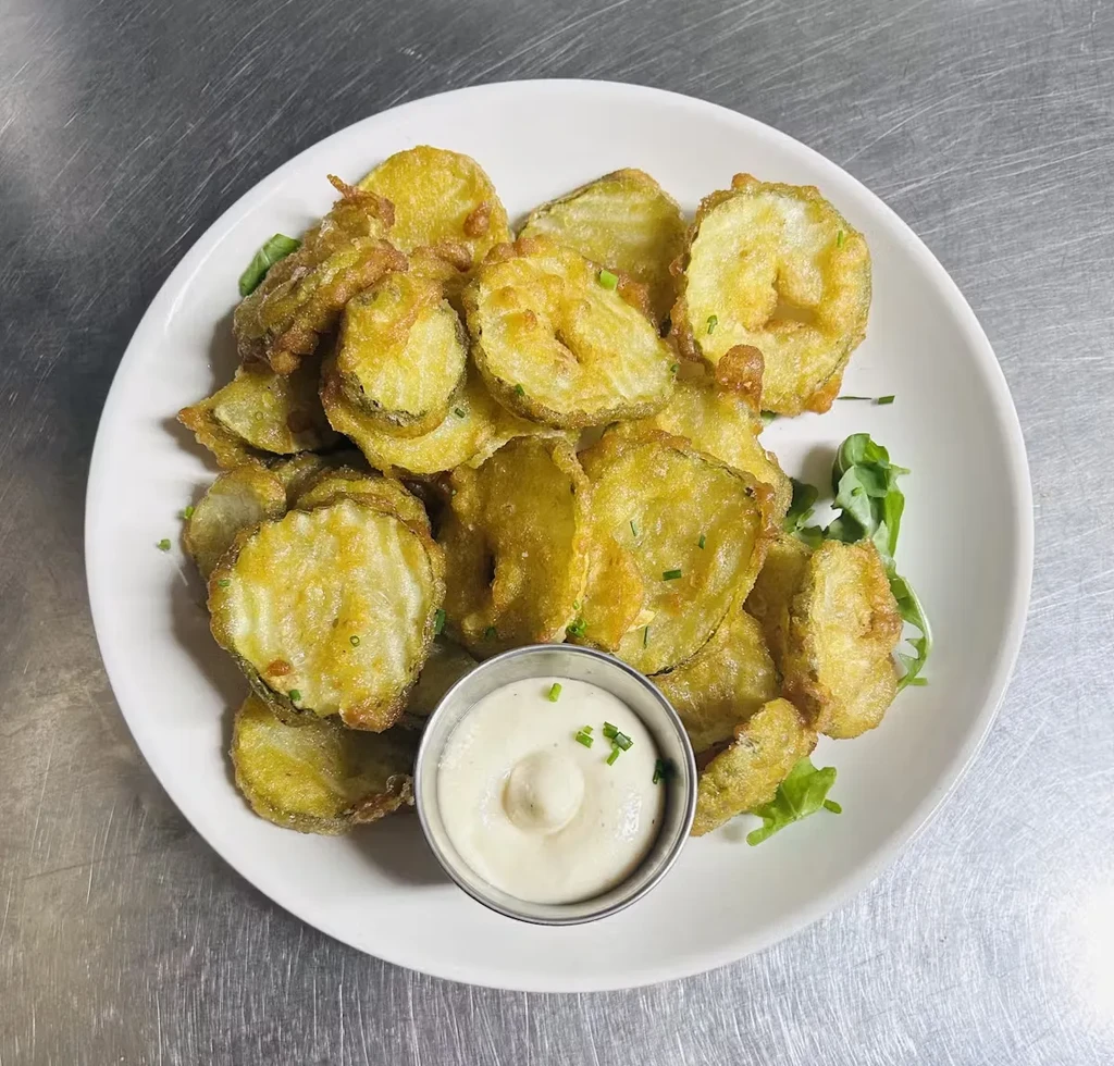 A white plate of fried pickles with dip and green garnish.