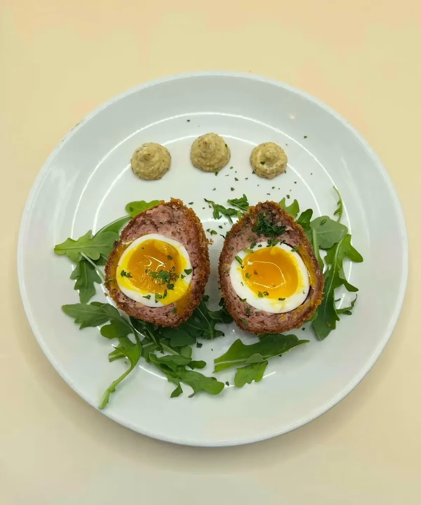 Two halved Scotch eggs with runny yolks, mustard, and arugula on a plate.