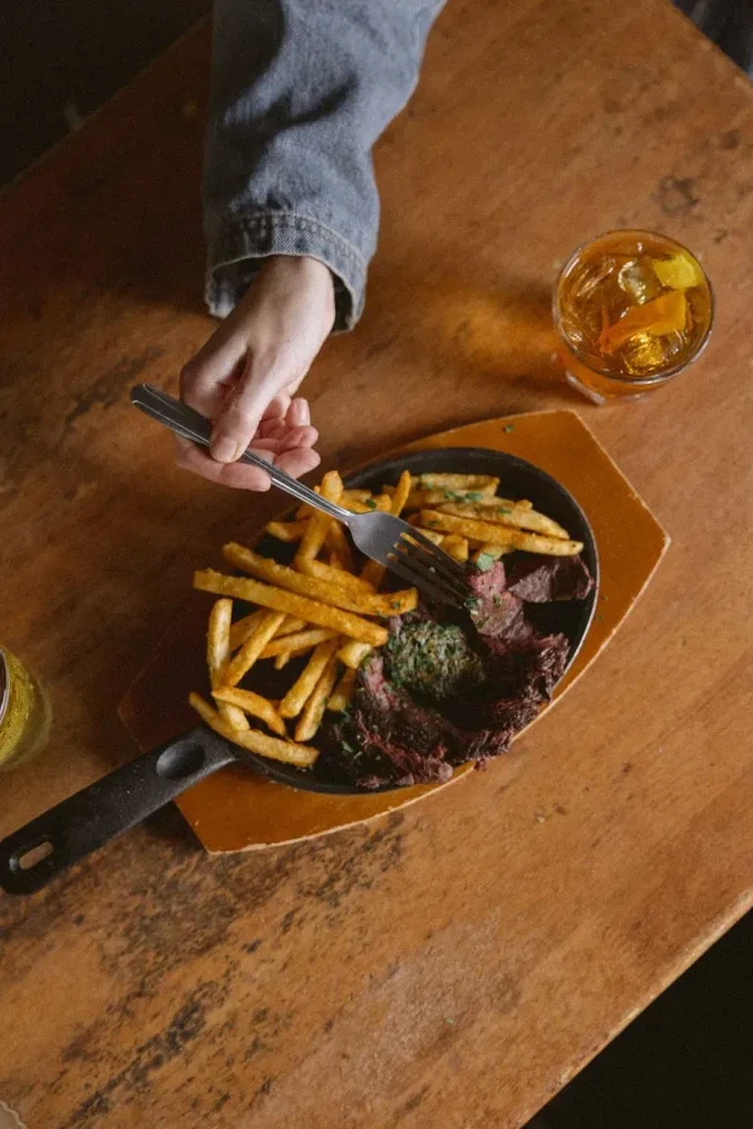A hand holding a fork in a black skillet with steak and fries.