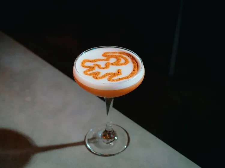 A cocktail with white foam topped with an orange powder design.