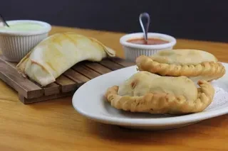 Three empanadas on plates with two dipping sauces on a wooden table.