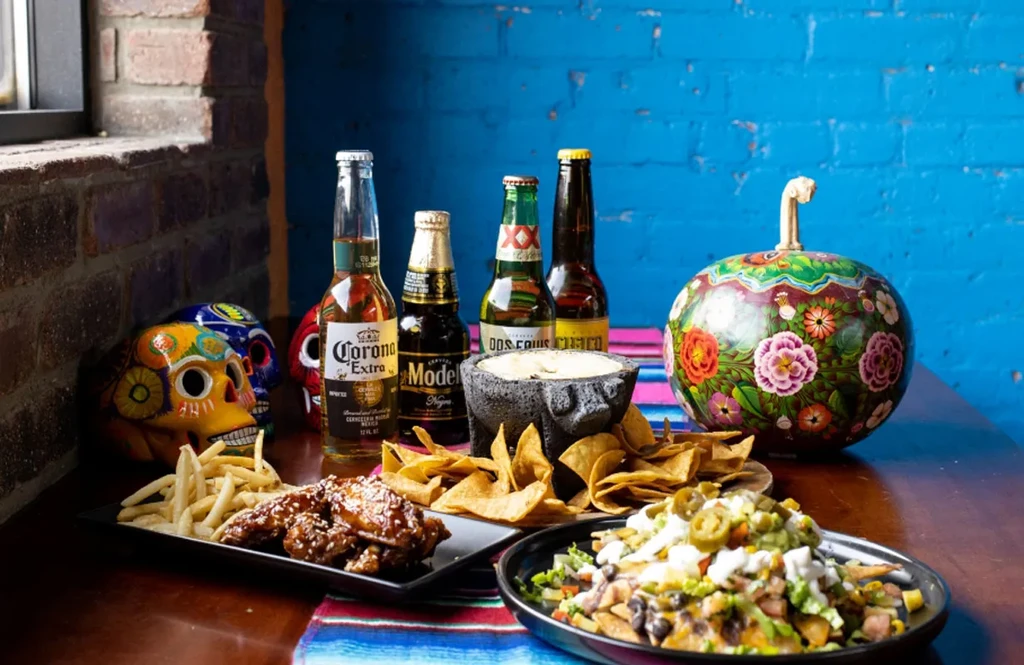 Mexican food spread with beer, nachos, chicken wings, fries, and decorative skulls.