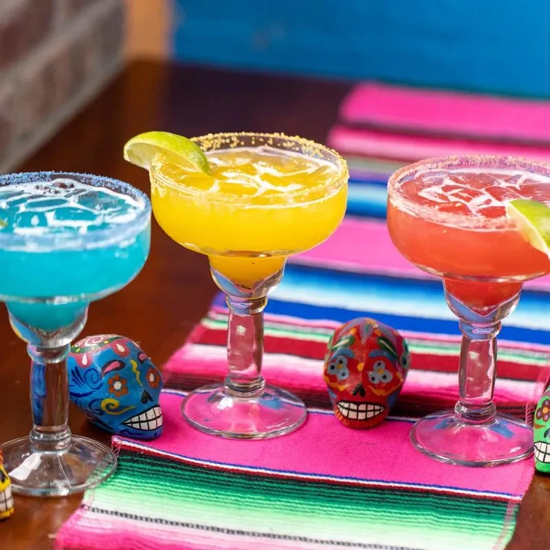 Three colorful margaritas sit on a vibrant striped serape with sugar skulls.