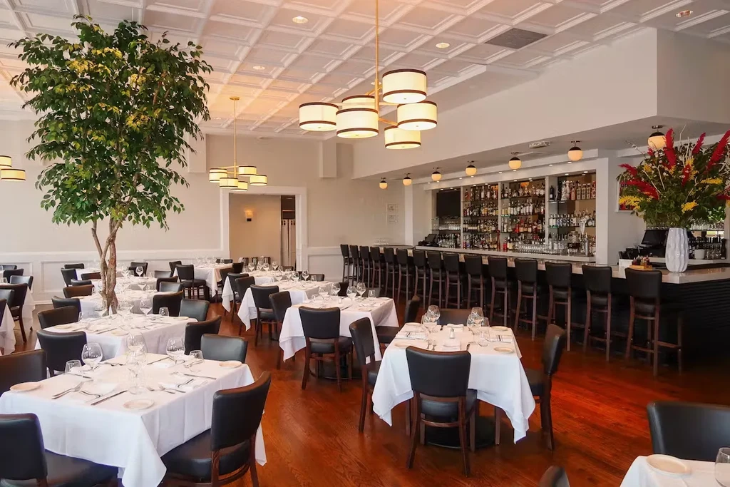 A restaurant dining room with white tablecloths, dark chairs, and a bar.