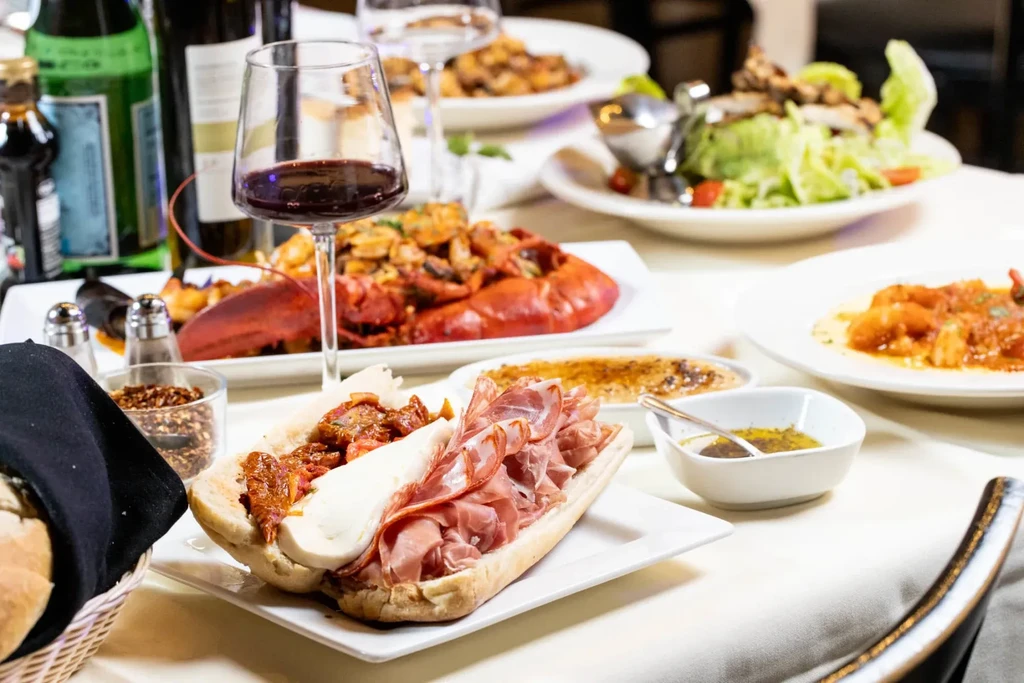 A table full of food, including lobster, a sub sandwich, and wine.