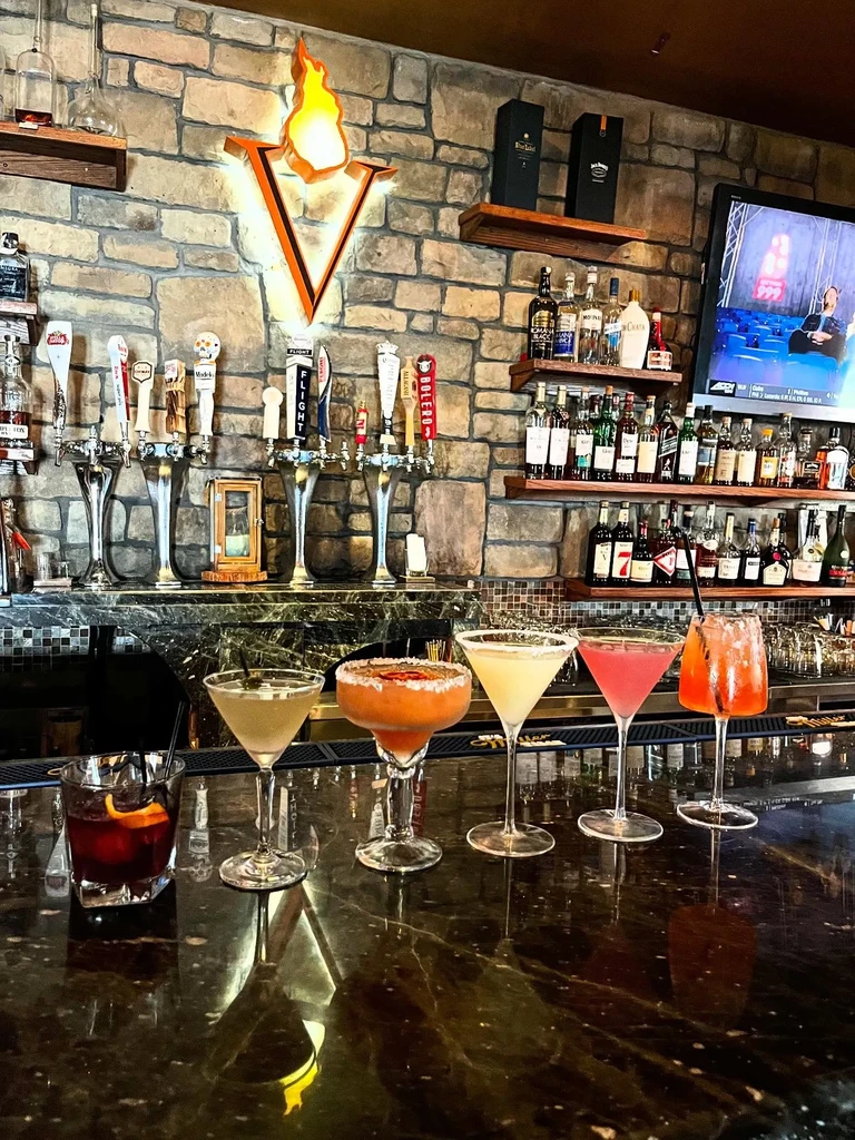 An illuminated "V" sign above a bar with cocktails on the counter.