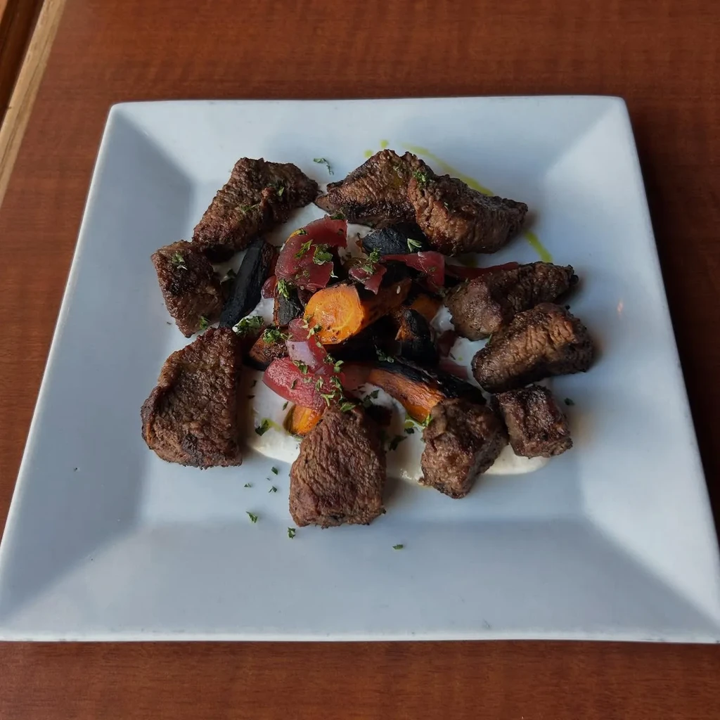 A white plate of cooked beef chunks, roasted vegetables, and white sauce.