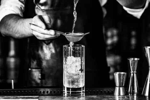 Black and white photo: Bartender straining a drink into a fluted glass with ice.