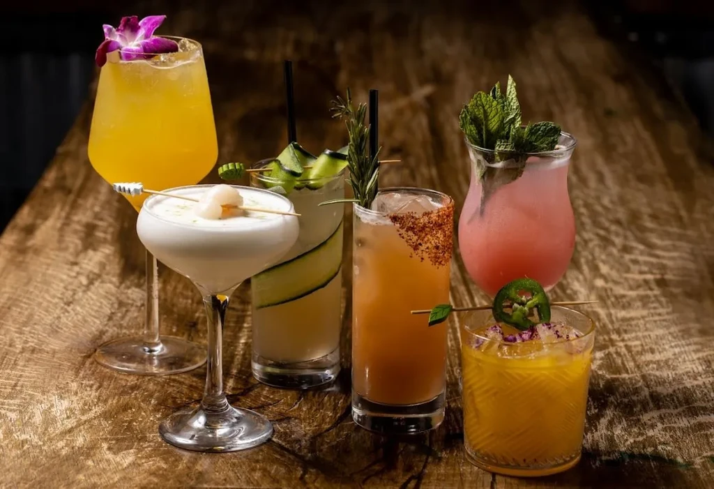 Six colorful cocktails, garnished with fruit, herbs, and spices, on a wooden table.