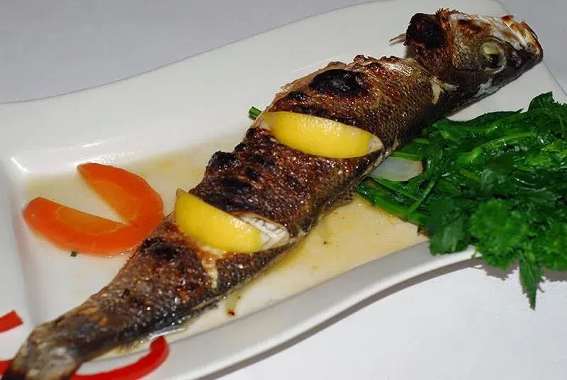 Grilled whole fish with lemon, greens, and tomato on a white plate.