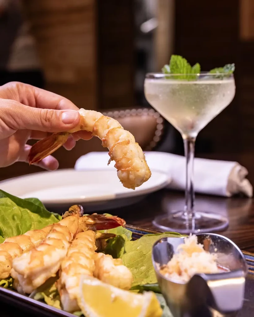 A hand holds a cooked shrimp over a plate with more shrimp and a drink.