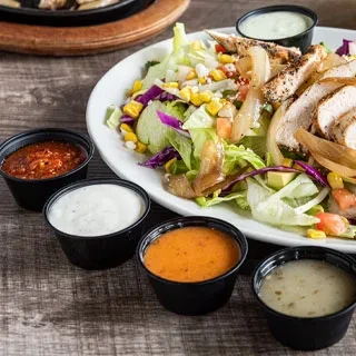 A plate of chicken salad with four bowls of sauce on a wooden table.