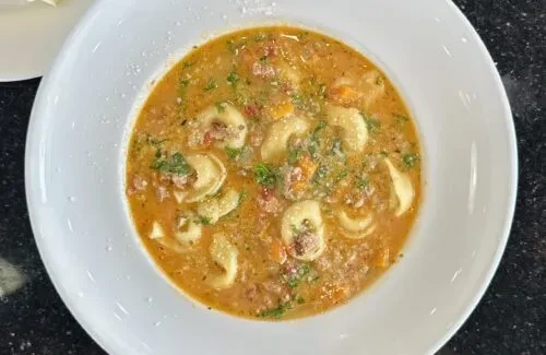 A white bowl of tortellini soup with meat, vegetables, and parmesan.