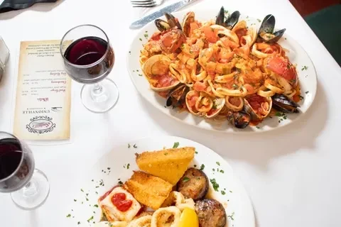 Two plates of seafood and two glasses of red wine on a white tablecloth.