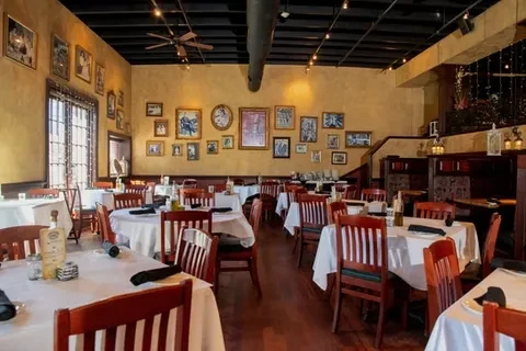 Empty restaurant dining room with yellow walls, framed art, and wooden chairs.