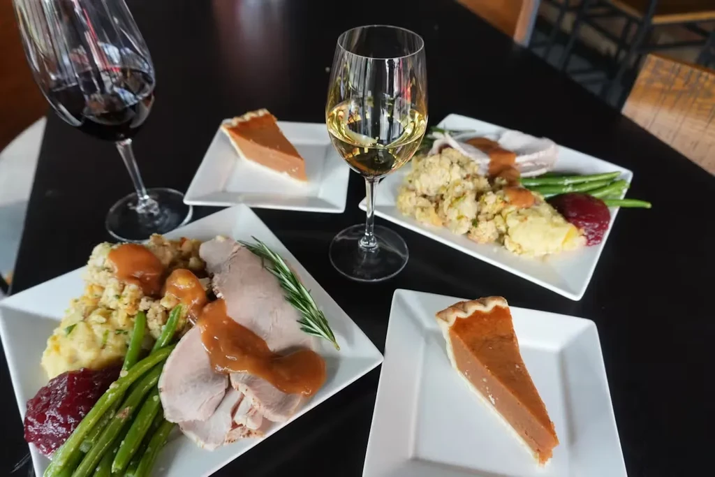Four plates with Thanksgiving turkey dinner, pumpkin pie, and two glasses of wine.