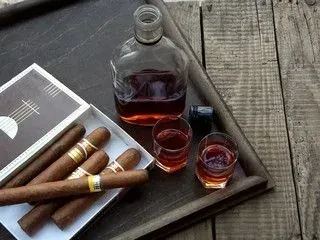 Amber spirit bottle, two glasses, and cigars on a wooden tray.