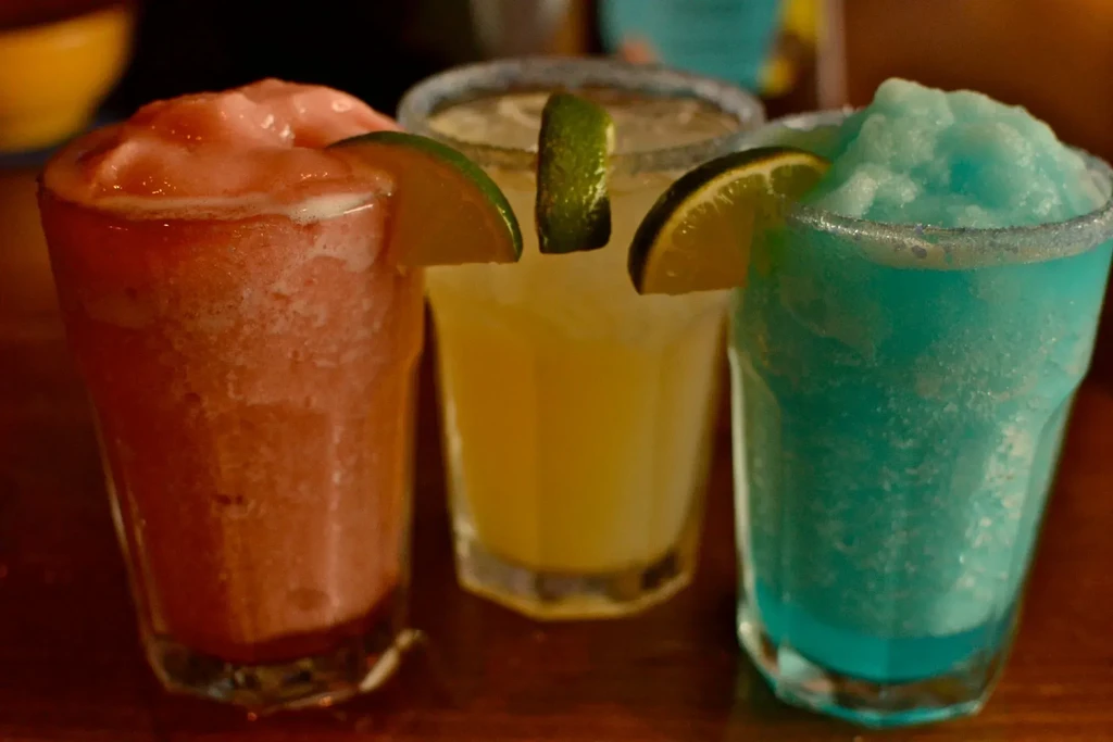 Three colorful frozen margaritas in glasses with lime wedges on a bar.