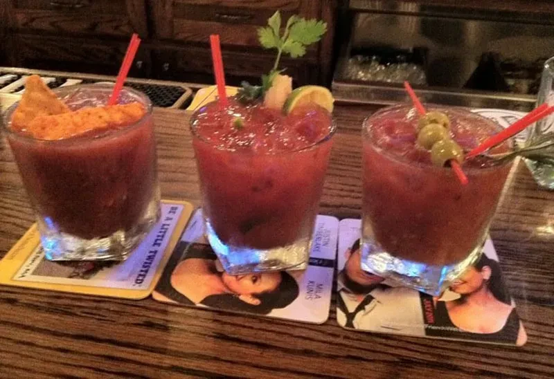Three Bloody Mary cocktails with various garnishes sit on a wooden bar.