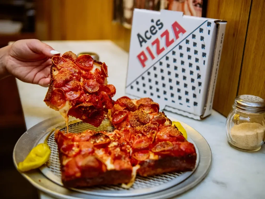 Hand holding a slice of square pepperoni pizza from Ace's Pizza.
