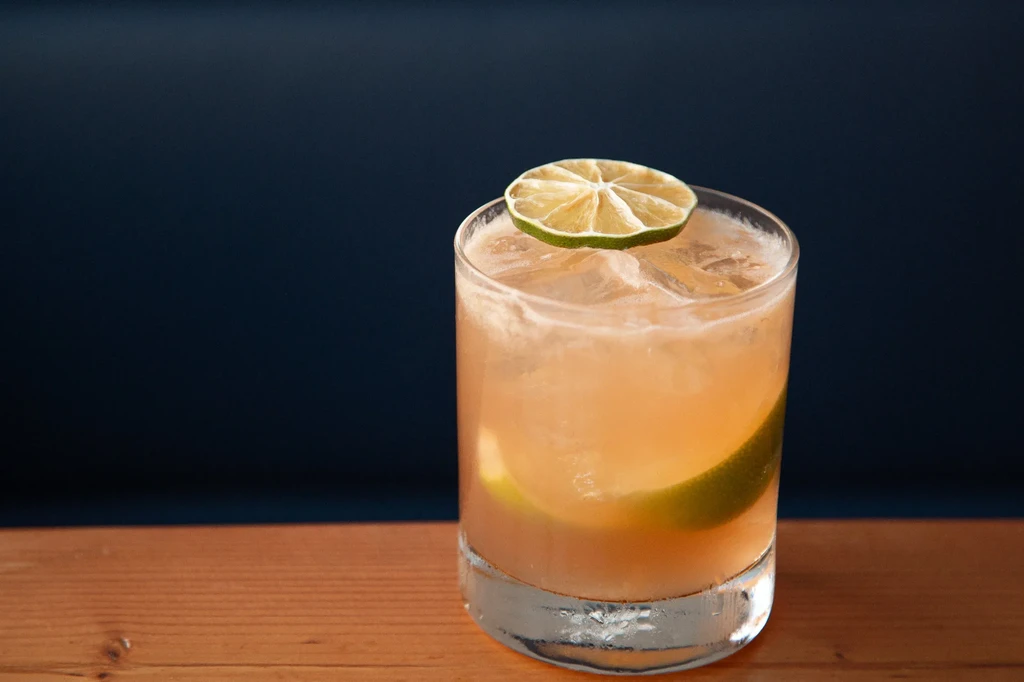 A glass of peach-colored cocktail with ice and lime slices.