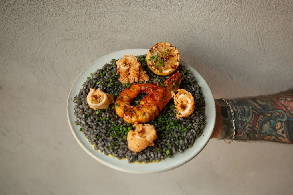 A tattooed arm holding a plate of squid ink risotto with prawns.