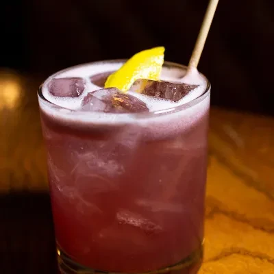 A red cocktail with ice, a lemon peel, and a straw.