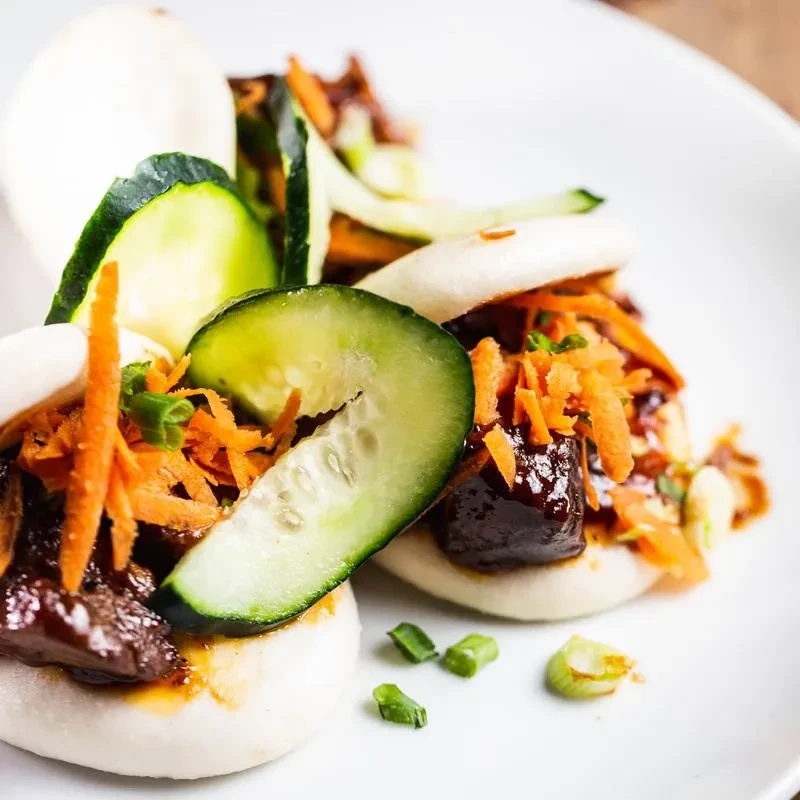 Two bao buns with meat, shredded carrots, and cucumber slices on a plate.