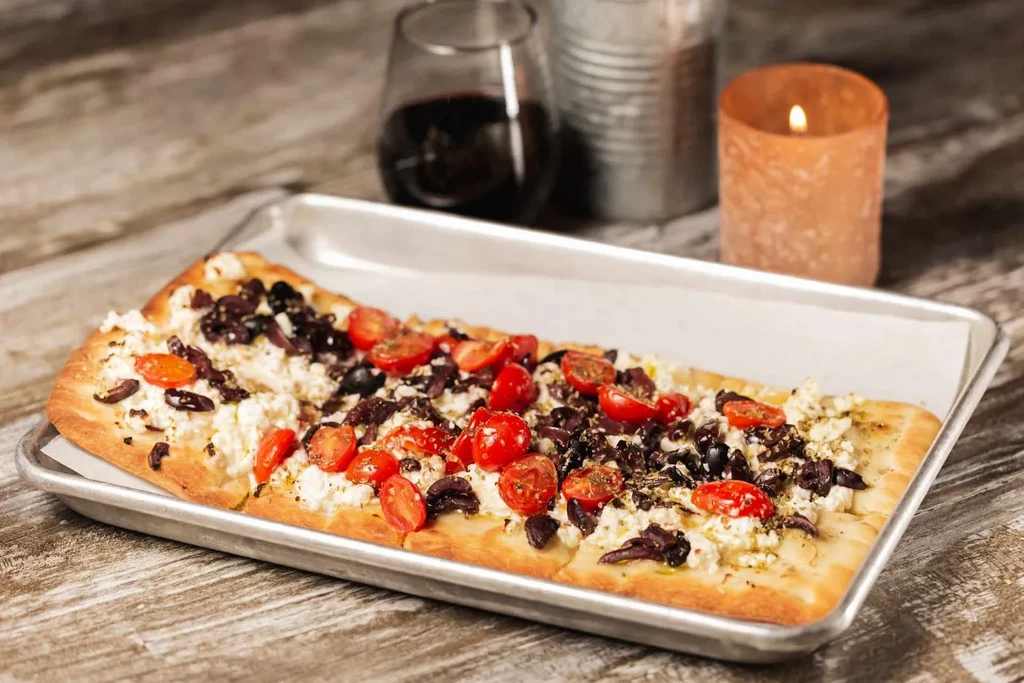 Flatbread with feta, olives, and tomatoes on a metal tray.