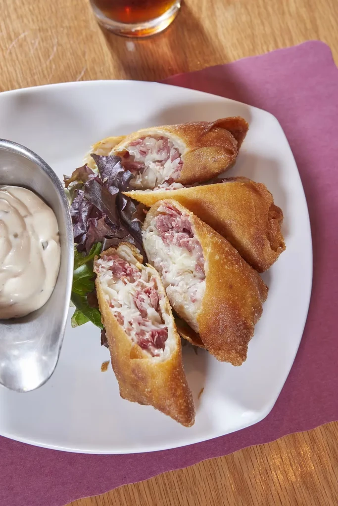 A plate of golden-brown reuben egg rolls with sauce and greens.