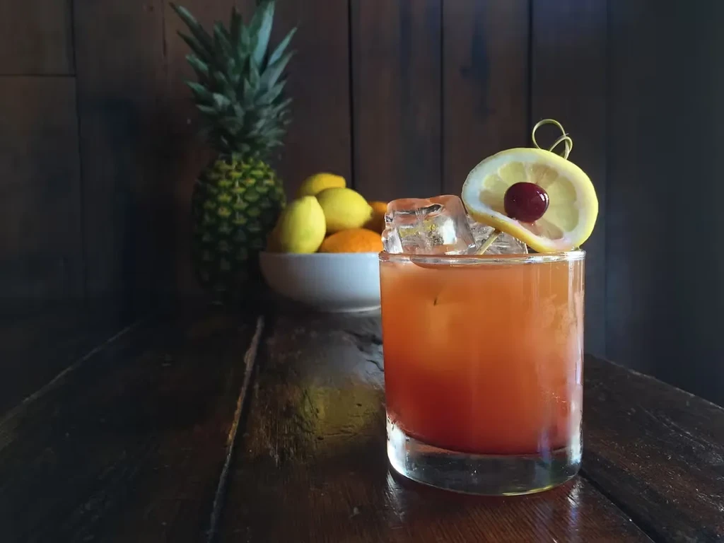 Cocktail with lemon and cherry garnish, fruit bowl, and pineapple.
