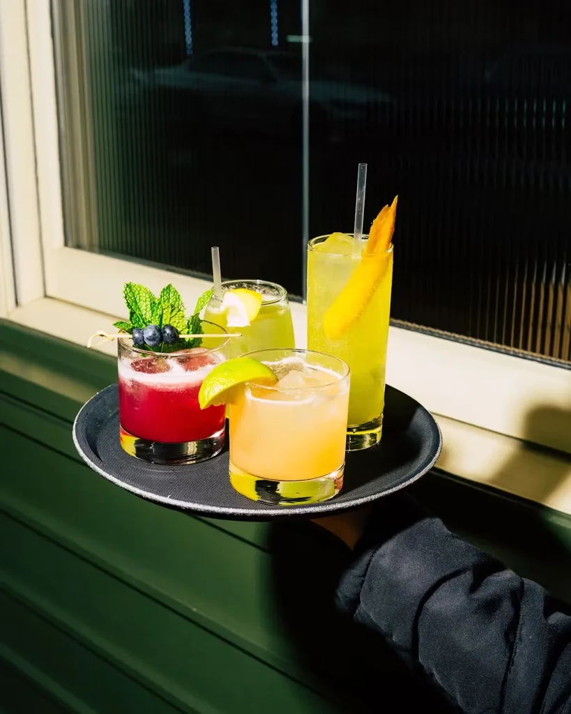 A person holding a black tray with four colorful cocktails.
