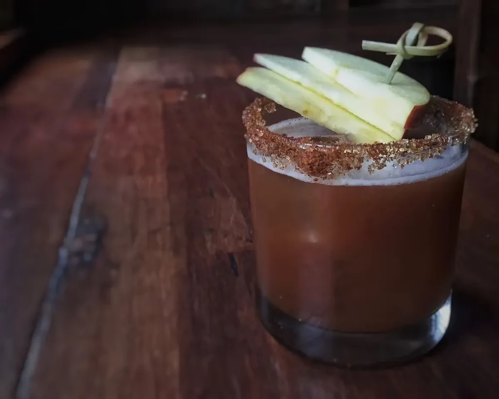 A brown cocktail with an apple slice on a wooden table.