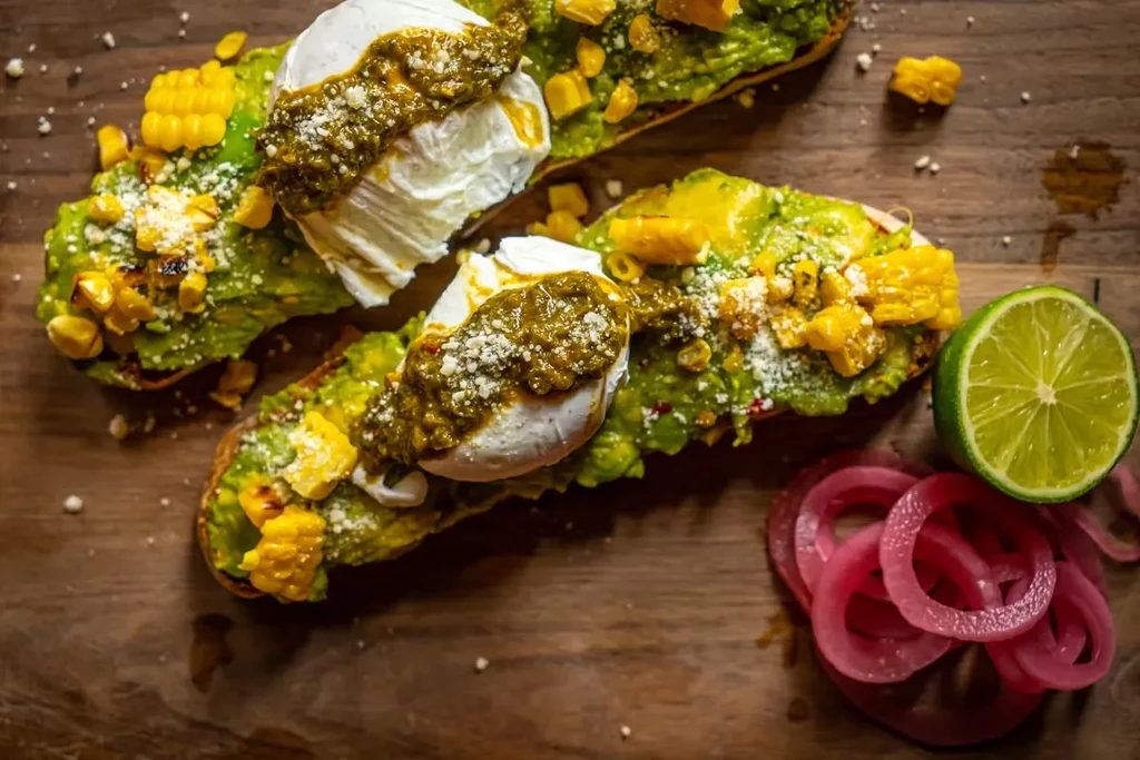 Two avocado toasts topped with poached eggs, corn, and green sauce.