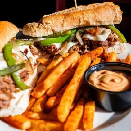 Two steak and cheese sandwiches with fries and dipping sauce.
