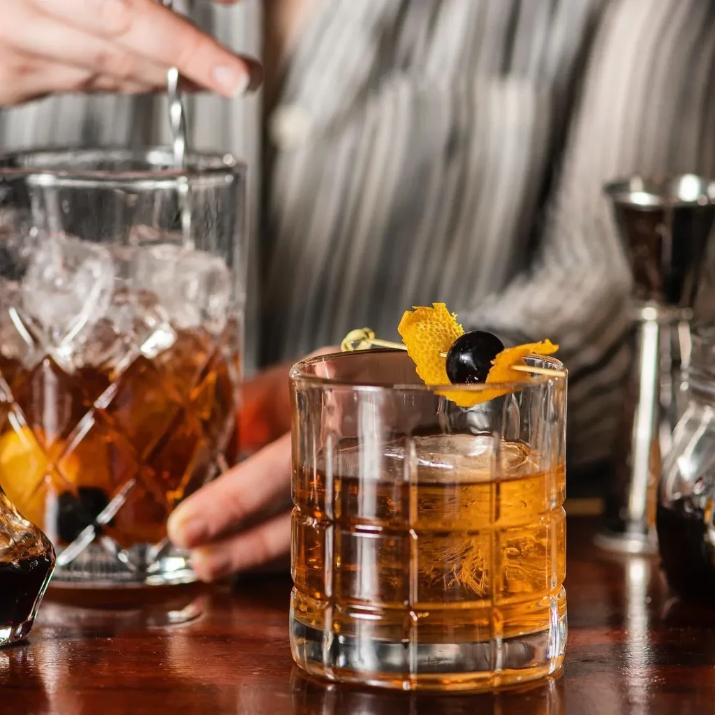 A bartender stirs cocktails with ice, garnished with orange peel and cherry.