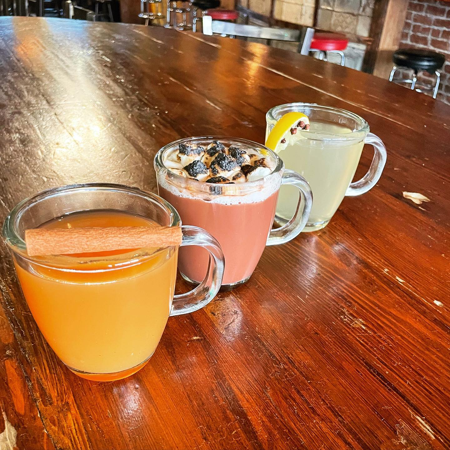 Three beverages in clear mugs with different garnishes on wood.