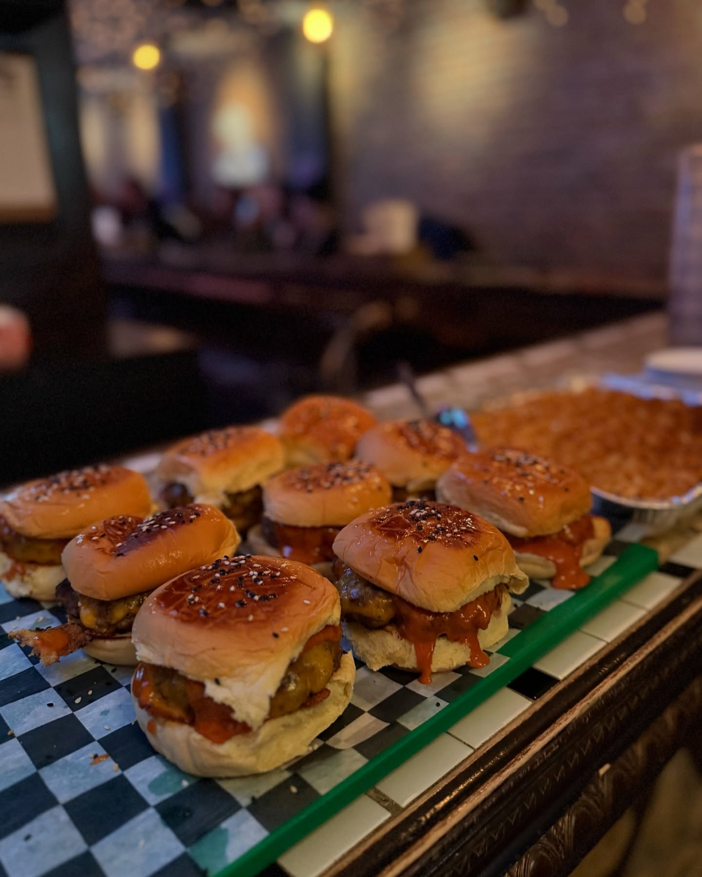 Multiple mini burgers with sauce and sesame seeds on a checkered tray.