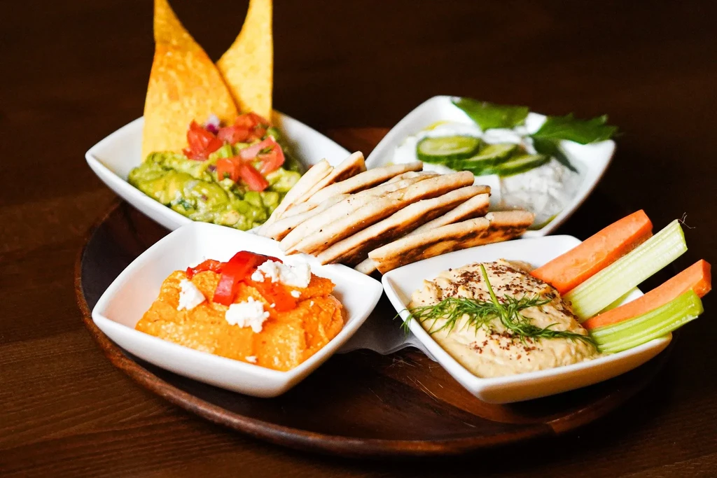 A wooden platter with hummus, guacamole, red pepper dip, tzatziki, and pita.