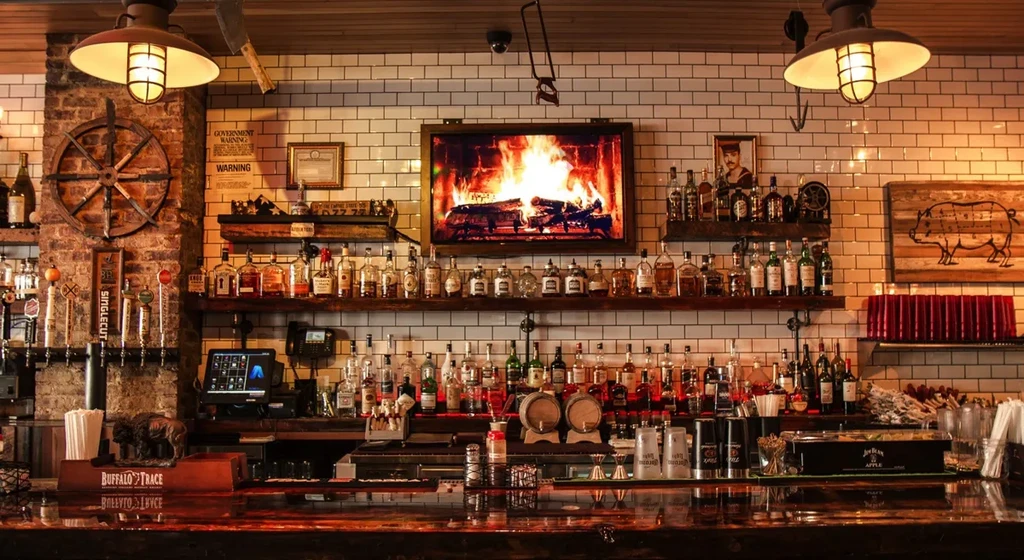 A fully stocked bar with a TV displaying a fireplace over a white tiled wall.